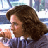Pixelated static image of a person with brown curly hair wearing a dark jacket over a light shirt, set against a blurred background with blue and white tones.