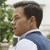 Pixelated profile view of a person with short dark hair wearing a white shirt and blue jacket, standing outdoors near green foliage.