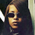 Pixelated close-up of a person with light brown hair wearing dark sunglasses and a dark jacket, set against a dark background with a light vertical edge on the right.