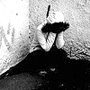Black-and-white pixelated image of a person sitting with knees drawn up, holding a dark object over their face, against a textured background.