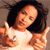 A pixelated image of a person with dark hair wearing a light-colored top, making a gesture with both hands raised near the shoulders.