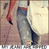 A person wearing light blue ripped jeans and a white top, holding a dark object in their right hand, with a white bag slung over their shoulder.