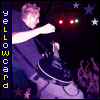 A person in a dark shirt plays an electric guitar under purple lighting with red and green stage lights, stars, and crowd silhouettes visible.