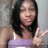 A person with long dark hair wears a light purple sleeveless top and makes a peace sign with their right hand against a blurred background.
