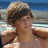 Pixelated image of a person with short dark hair wearing a light-colored sleeveless top, standing against a blurred beach background with blue sky and water.