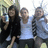 Three people pose outdoors with one holding a blue bottle; the rightmost person's arm position changes between frames.