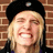 Pixelated close-up of a person with blonde hair wearing a black cap, smiling broadly against a brick wall background.