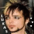 Pixelated close-up of a person with light brown hair and dark roots, wearing a black choker necklace against a dark background.