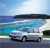 Pixelated static image of a white sedan on a road with blue water and green grass in the background under a light blue sky.