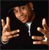 Pixelated image of a person in a dark suit with a white shirt, making a gesture with both hands raised and fingers spread against a black background.