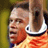 Pixelated static image of a person in an orange sports jersey with white trim and dark collar, wearing a headband or cap, against a blurred green background.