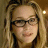 A pixelated close-up of a person with light brown hair and dark-framed glasses, wearing a dark top against a warm-toned background.