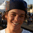 Pixelated close-up of a person with light skin wearing a dark cap, smiling with eyes partially closed against a blurred outdoor background.