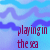 Pixelated static image with blue and purple gradient background featuring wavy cloud-like shapes and maroon text reading "playing in the sea" at bottom right.