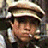 A pixelated close-up of a person wearing a dark cap with a light band, light-colored shirt, and dark jacket, with a blurred background.