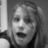 Grayscale pixelated image of a person with light hair, mouth open in mid-expression, wearing dark clothing against a neutral background.