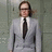 Pixelated static image of a person in a light gray suit with a white shirt and dark tie against a muted green background.