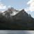 Pixelated landscape showing dark mountains under a light blue sky with scattered white clouds, reflected in a calm body of water below.