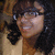 A person with dark curly hair and glasses smiles in a static image, wearing a light-colored top against a blurred background.