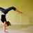 Pixelated figure in dark blue top and black shorts performing a handstand against a gradient yellow background.
