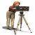 A woman with orange hair and red top stands behind a tripod-mounted camera, leaning forward to view through the lens.