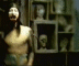 A shirtless figure with long dark hair stands in a dimly lit room filled with framed busts and skulls, arms extended outward.