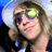 Pixelated static image of a person with brown hair holding a microphone, wearing dark clothing and green-tinted glasses against a blue background.