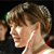 Pixelated close-up of a person's head and neck in profile, showing light brown hair, a visible ear, and a white strap across the neck against a dark background.