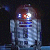 Pixelated static image of a humanoid robot with blue and silver coloring, red accents on the head, and glowing white eyes against a dark background.