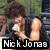 Nick Jonas holds a microphone, wearing a black sleeveless shirt with green foliage and dark structures in the background.