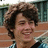A person with curly brown hair wearing a blue and white checkered shirt smiles against an outdoor background.