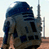 Pixelated static image of a blue and gray robot-like figure with dark accents against a light background, appearing to be a low-resolution digital icon.