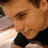 Pixelated close-up of a person's face with light brown hair, dark eyes, and a black garment visible at the bottom, set against a blurred background.
