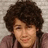 Pixelated close-up of a person with voluminous dark curly hair and light skin wearing a brown garment against a neutral background.