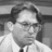 Black-and-white pixelated image of a person with short dark hair, wearing glasses and a light-colored shirt against a blurred background.
