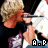 A pixelated image of a person with light brown hair holding a silver microphone, wearing a dark top and patterned shirt, with 'A.P' text at the bottom.