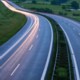 Static image of a winding road with light trails at dusk or dawn, surrounded by greenery and trees.