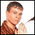 Pixelated static image of a person with short brown hair wearing a brown patterned shirt and dark necklace, holding an object against white background.