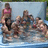 Group of people in a pool with one person standing and others seated, wearing swimwear.