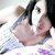 Pixelated image of a person with dark hair tied in a high ponytail, wearing a light-colored top and holding a small brown object near their face.
