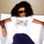 A person with voluminous black hair wears a white T-shirt featuring blue and pink graphic elements, dark sunglasses, and dark pants against a light brown background.