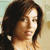 A pixelated close-up of a person with long dark brown hair, light skin, and a neutral expression, wearing a light-colored top against a blurred background.