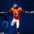 Pixelated image of a football player in an orange jersey with white pants, arms outstretched against a dark blue background.