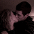 Two people in a dimly lit room, one with short dark hair and the other with long light hair, sharing a kiss with closed eyes.