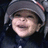 Pixelated close-up of a smiling person wearing a gray beanie with red stripe, dark clothing against black background.
