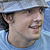 Static close-up of a person's head and shoulders wearing a light blue cap against a blurred background with visible pixelation.
