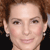 Pixelated close-up of a person's face with light skin, brown eyes, and curly brown hair, wearing small earrings and a dark top.