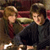 Two figures in dark clothing stand close together against a blurred background with warm tones and indistinct shapes.