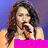 A woman with long brown hair holds a microphone, wearing a pink top against a blue background.