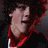 Pixelated close-up of a person's face with light skin tone, dark hair, and red clothing against a dark background.